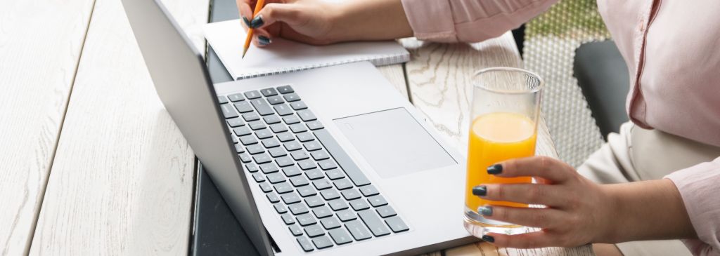 Close up portrait of a young woman working on laptop and writing, holding a glass of orange juice.