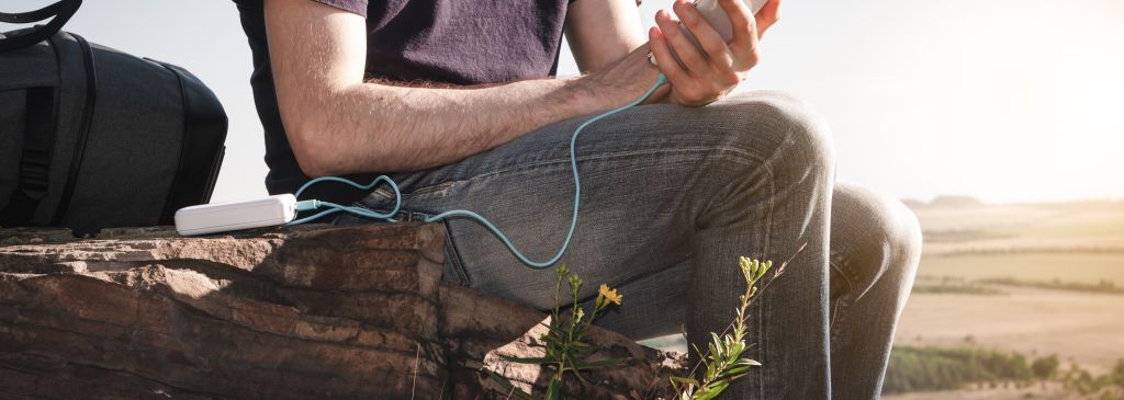 Man on a hike uses smartphone while charging from the power bank on the rock at dawn. Healthy lifestyle and communication.